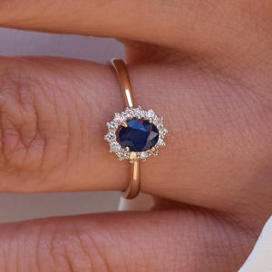 Könnte beinhalten: Ein goldener Ring mit einem dunkelblauen, oval geschliffenen Edelstein, umgeben von kleineren, klaren Steinen. Das Design des Rings steht im Mittelpunkt, vor einem verschwommenen Hintergrund.