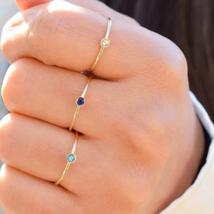 May include: Three gold rings with small gemstones. The rings are stacked on a person's finger. The top ring has a clear gemstone, the middle ring has a dark blue gemstone, and the bottom ring has a light blue gemstone.