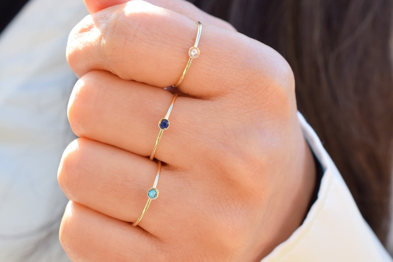May include: Three gold rings with small gemstones. The rings are stacked on a person's finger. The top ring has a clear gemstone, the middle ring has a dark blue gemstone, and the bottom ring has a light blue gemstone.