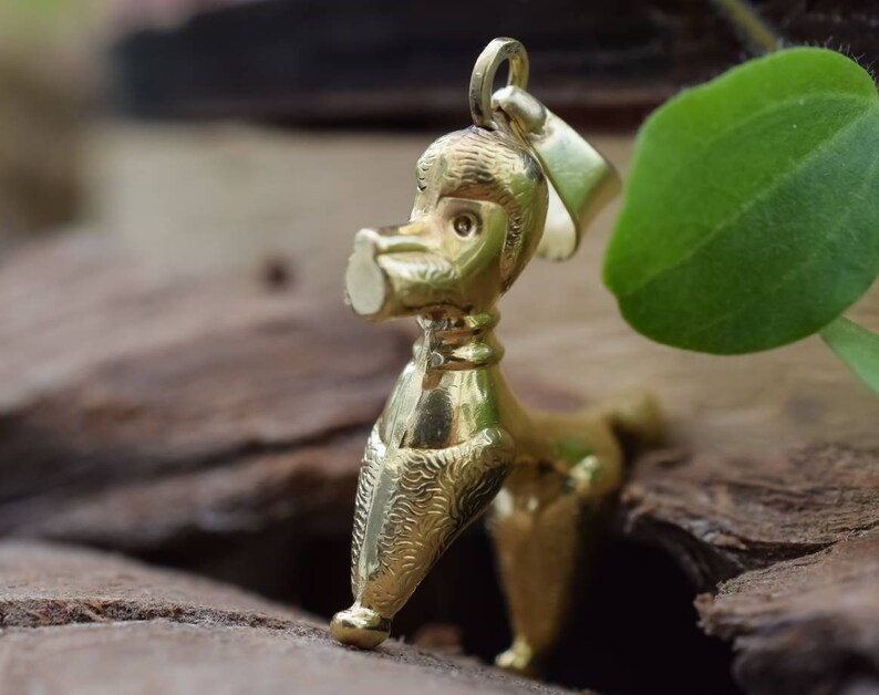 Puede incluir: Un colgante de caniche de color dorado. El caniche est&aacute; de pie sobre sus cuatro patas con la cabeza inclinada hacia un lado. El colgante tiene un dise&ntilde;o detallado y un acabado brillante.