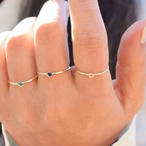 May include: Three gold rings with small gemstones. The rings are on a person's finger. The gemstones are blue, clear, and green.