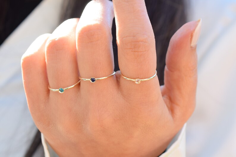 May include: Three gold rings with small gemstones. The rings are on a person's finger. The gemstones are blue, clear, and green.
