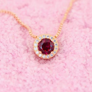 Può includere: Una collana d'oro con un ciondolo di pietra rossa circondato da un'aureola di piccole pietre bianche.