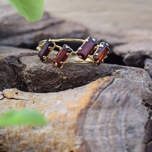 May include: A gold ring with five rectangular red gemstones set in a delicate band. The ring is resting on a piece of weathered wood.