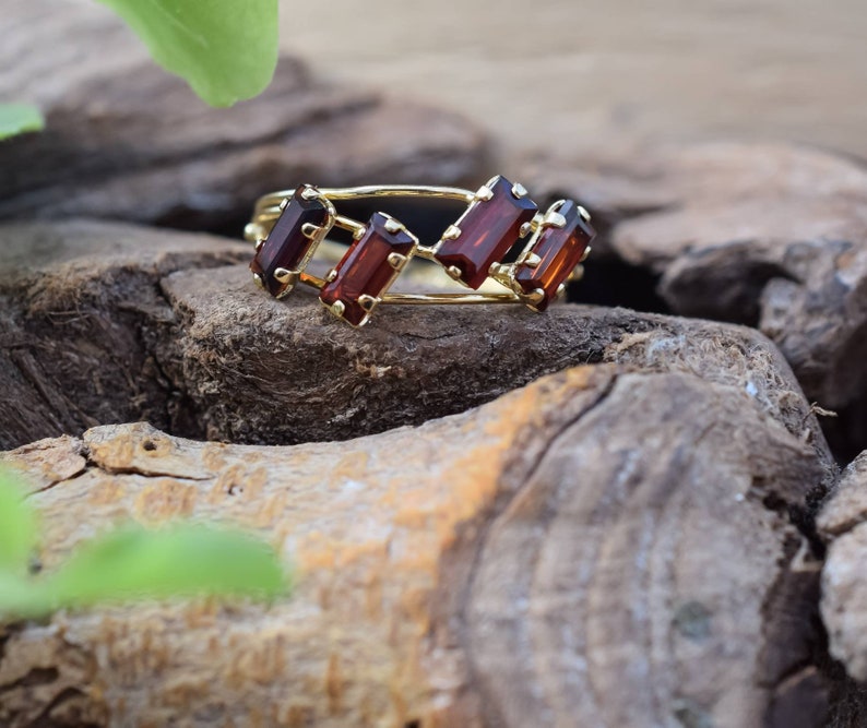 May include: A gold ring with five rectangular red gemstones set in a delicate band. The ring is resting on a piece of weathered wood.