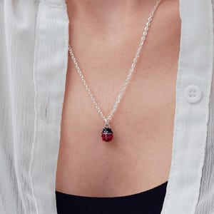 May include: A silver chain necklace featuring a small, round ladybug pendant. The pendant is red with black spots and a silver top. The necklace is worn with a white button-down shirt over a black top, highlighting the contrast of the jewelry.