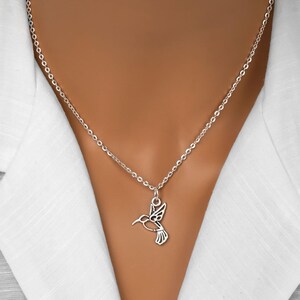 May include: A silver-toned necklace featuring a delicate, openwork hummingbird pendant suspended from a simple link chain. The pendant is shaped like a stylized bird in flight, showcasing clean lines and a polished metallic finish against a white shirt.