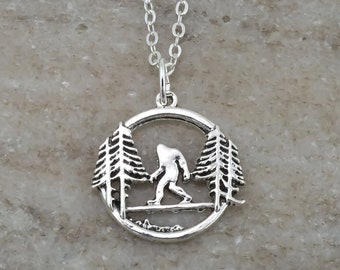 Puede incluir: Un collar de tono plateado que presenta un colgante circular con un diseño recortado de una figura de Bigfoot caminando entre dos pinos. El colgante está unido a una delicada cadena de eslabones de tono plateado, creando una pieza de joyería.