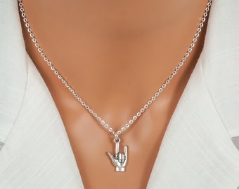 May include: A silver-toned necklace featuring a pendant shaped like a hand making the I love you sign in American Sign Language. The pendant hangs from a delicate silver-toned chain that rests against a white collared shirt.