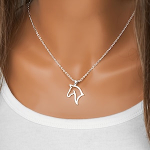 May include: A silver-toned necklace featuring a delicate chain and a minimalist pendant shaped like the outline of a horse head. The pendant is polished and hangs centered on the chain, which rests against a plain white fabric surface.
