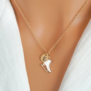 May include: A gold-toned chain necklace featuring two charms: a small circular gold disc engraved with the capital letter E and a white ice skate charm with gold-toned metal accents, suspended from a delicate gold-toned link chain.