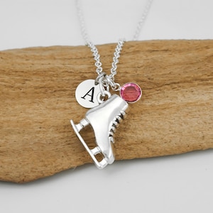 May include: A silver-toned necklace featuring a pendant shaped like an ice skate, a small circular charm engraved with the letter A, and a round pink crystal accent. The charms are suspended from a delicate silver-toned chain link necklace.