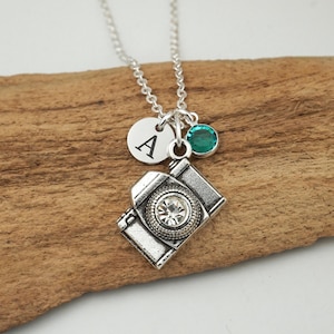 May include: A silver-toned necklace featuring a camera-shaped pendant with a clear crystal lens, a small circular charm engraved with the letter A, and a single round teal-colored crystal bead suspended from a delicate silver-toned metal chain.