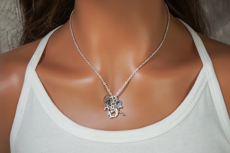 Puede incluir: Un collar de cadena plateada con tres dijes: un disco circular grabado con la letra A, una peque&ntilde;a piedra preciosa azul claro facetada y una figura de caballo plateada detallada, sobre una camiseta sin mangas blanca.