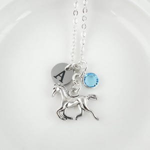 Puede incluir: Un collar de tono plateado que presenta un dije de caballo met&aacute;lico, un colgante circular grabado con la letra A y un peque&ntilde;o dije de cristal redondo de color azul claro. Los dijes cuelgan de una delicada cadena de eslabones plateada.