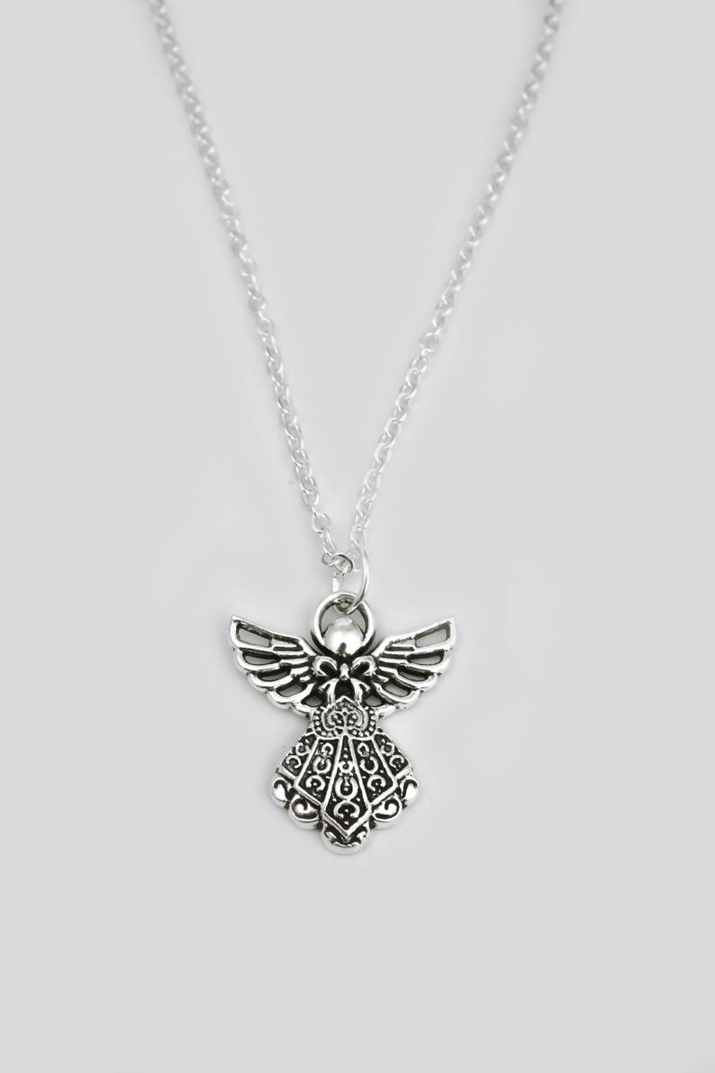 May include: A silver-toned metal necklace featuring an angel-shaped pendant. The pendant displays intricate cut-out detailing on the wings and a decorative filigree pattern on the dress, suspended from a simple, thin silver-toned link chain.
