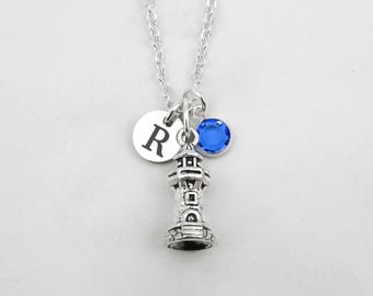 May include: A silver-toned necklace featuring a lighthouse charm, a circular disc charm engraved with the letter R, and a small round blue crystal charm. The charms are suspended from a delicate silver-toned metal chain link necklace.