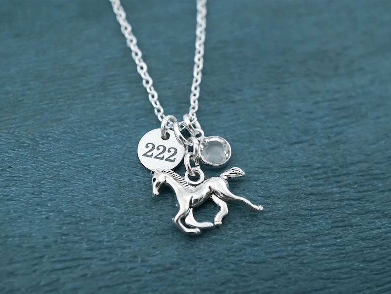 Puede incluir: Un collar de tono plateado con una cadena fina y tres dijes: un colgante circular grabado con el n&uacute;mero 222, una peque&ntilde;a cuenta de cristal transparente y un dije detallado de un caballo en posici&oacute;n de galope, unidos por una argolla.