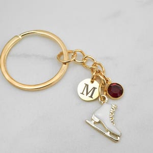 May include: A gold-toned metal keychain featuring a circular ring attached to a chain with three charms: a round gold disc engraved with the letter M, a white enamel ice skate charm with gold details, and a small round dark red crystal bead.