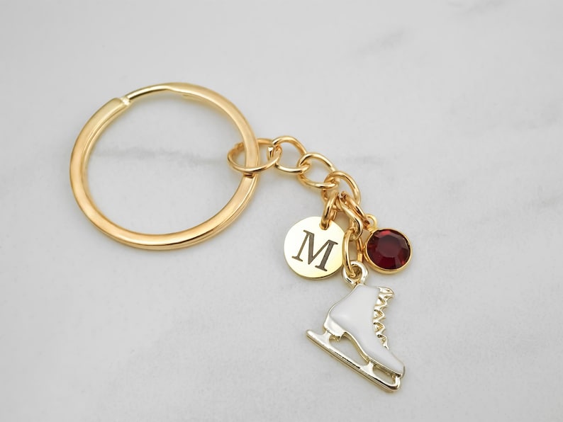 May include: A gold-toned metal keychain featuring a circular ring attached to a chain with three charms: a round gold disc engraved with the letter M, a white enamel ice skate charm with gold details, and a small round dark red crystal bead.