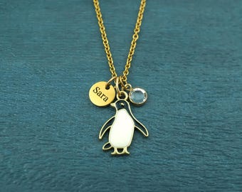 May include: A gold-toned necklace featuring a central penguin pendant with black and white enamel detailing. Attached to the chain are two additional charms: a small round gold disc engraved with the name Sara and a clear round crystal.