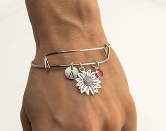 May include: A silver-toned expandable bangle bracelet featuring three dangling charms: a circular disc engraved with the letter A, a detailed sunflower charm with textured petals, and a small, round, translucent pink gemstone charm.