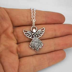 May include: A silver-toned metal angel pendant necklace featuring an intricate filigree pattern on the wings and dress. The angel figure is suspended from a delicate silver-coloured link chain, showcasing a polished finish with dark recessed details.