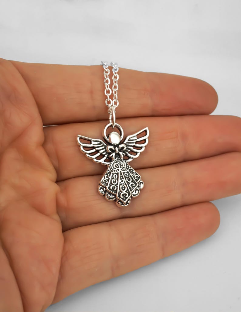 May include: A silver-toned metal angel pendant necklace featuring an intricate filigree pattern on the wings and dress. The angel figure is suspended from a delicate silver-coloured link chain, showcasing a polished finish with dark recessed details.