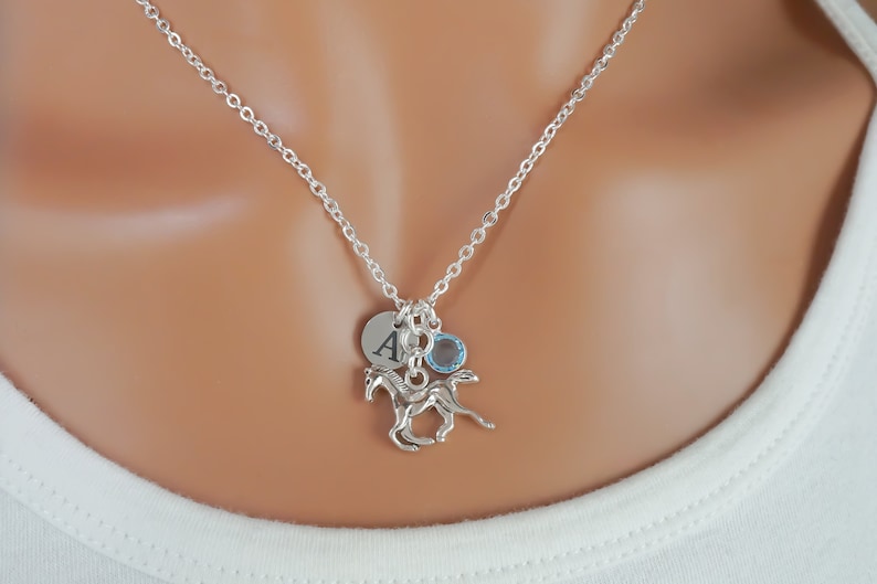 Puede incluir: Un collar de tono plateado con una cadena fina y tres dijes: un disco circular grabado con la letra A, una peque&ntilde;a piedra preciosa redonda de color azul claro y un dije plateado detallado de un caballo corriendo.