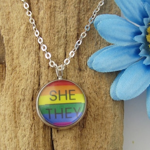 May include: A silver-toned pendant necklace featuring a circular charm with a rainbow-colored background and the black text SHE THEY printed on the front. The pendant hangs from a delicate silver-toned metal chain with small, uniform oval links.
