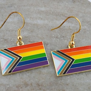 May include: A pair of gold-coloured earrings featuring a rectangular design with the Progress Pride flag. The flag design includes the traditional rainbow colours, along with a chevron of colours representing marginalised communities.