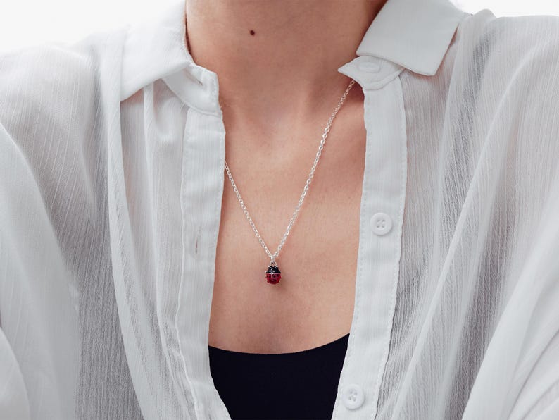 May include: A silver chain necklace featuring a small, round ladybug pendant. The pendant is red with black spots and a silver top. The necklace is worn with a white button-down shirt over a black top, highlighting the contrast of the jewelry.