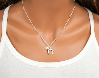 May include: A silver-toned necklace featuring a delicate chain with two charms: a small circular disc engraved with the letter E and a horseshoe charm with small circular indentations along its curved surface.