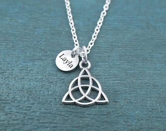 May include: A silver-toned necklace featuring a delicate chain, a circular charm engraved with the name Layla, and a triquetra pendant. The metal components have a polished finish, and the charms are suspended from a single jump ring on the chain.