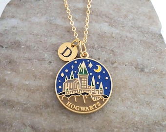 May include: A gold-tone necklace featuring a round pendant with a blue enamel background depicting a castle, a yellow crescent moon, and stars. The word HOGWARTS is printed at the bottom. A small circular charm with the letter D is attached.