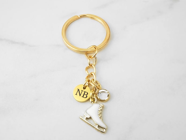 May include: A gold-toned metal keychain featuring a circular ring, a small round charm engraved with the letters NB, a clear faceted crystal bead, and a white enamel ice skate charm with gold-toned details attached by a short chain link.