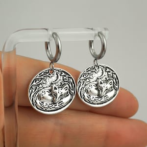 May include: A pair of silver-toned hoop earrings featuring circular charms. Each charm displays a detailed embossed design of a fox curled up in a forest setting under a crescent moon. The metal has an oxidized finish to highlight the textures.