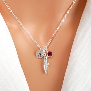 May include: A silver-toned necklace featuring a delicate chain with three charms: a circular pendant engraved with the letter A, a ballet slipper charm, and a small, round, deep red gemstone set in a silver-toned bezel.