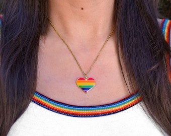 May include: A gold-toned chain necklace featuring a heart-shaped pendant with horizontal stripes in red, orange, yellow, green, blue, and purple enamel. The pendant hangs from a thin metal chain against a white top with rainbow trim.