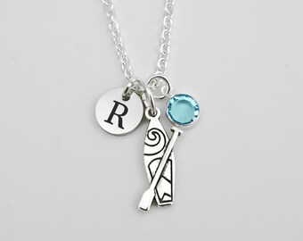 May include: A silver-toned necklace featuring three charms: a circular disc engraved with the letter R, a detailed kayak and paddle charm, and a small, round, light blue crystal stone. The charms hang from a delicate silver-toned chain link.
