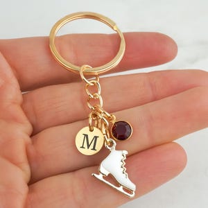 May include: A gold-toned metal keychain featuring a circular split ring, a small round charm engraved with the letter M, a white enamel ice skate charm, and a single round dark red crystal charm, all connected by a small gold-toned chain link.