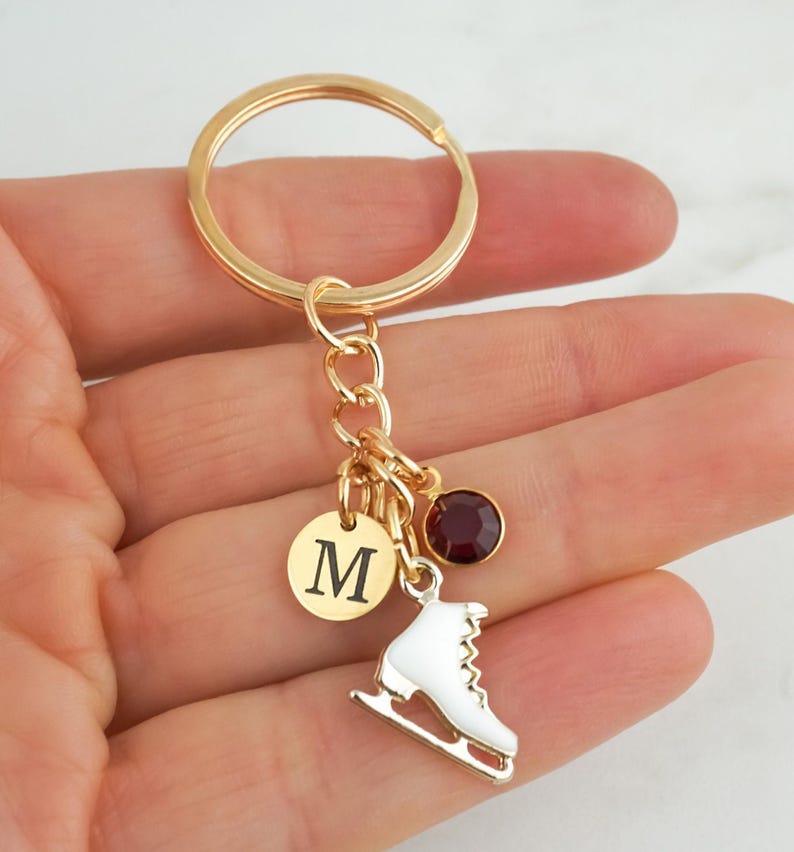 May include: A gold-toned metal keychain featuring a circular split ring, a small round charm engraved with the letter M, a white enamel ice skate charm, and a single round dark red crystal charm, all connected by a small gold-toned chain link.