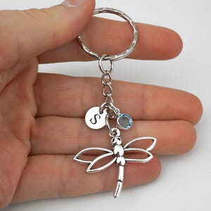 May include: A silver-toned metal keychain featuring a dragonfly charm, a small circular disc engraved with the letter S, and a single round light blue crystal charm attached to a split ring by a short silver chain link connector.