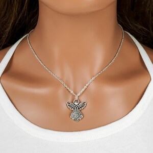 May include: A silver-toned necklace featuring an angel pendant with detailed wings and a textured dress design. The pendant hangs from a simple silver-toned chain that rests against the neck, showcasing a metallic finish and intricate metalwork.