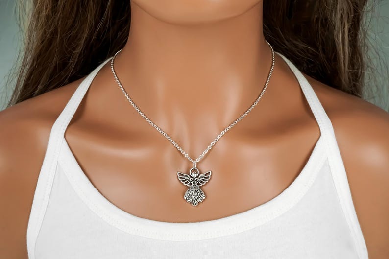 May include: A silver-toned necklace featuring an angel pendant with detailed wings and a textured dress design. The pendant hangs from a simple silver-toned chain that rests against the neck, showcasing a metallic finish and intricate metalwork.