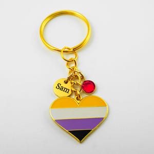 May include: A gold-colored keychain with a heart-shaped charm featuring the non-binary pride flag design. The keychain includes a small gold disc engraved with the name "Sam" and a red gem. The heart charm is yellow, white, purple, black, and gold.