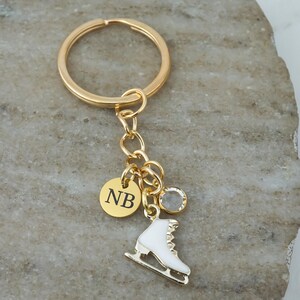 May include: A gold-toned metal keychain featuring a circular ring, a small round charm engraved with the letters NB, a clear crystal bead, and a white enamel ice skate charm. The components are connected by a gold-toned chain link assembly.
