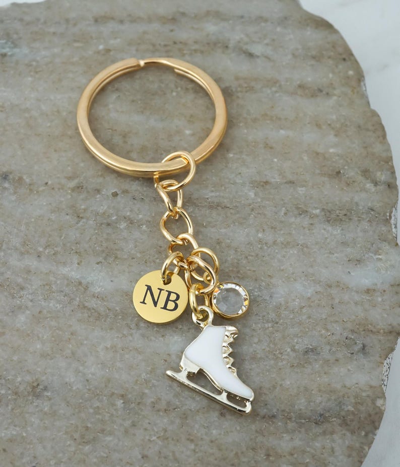 May include: A gold-toned metal keychain featuring a circular ring, a small round charm engraved with the letters NB, a clear crystal bead, and a white enamel ice skate charm. The components are connected by a gold-toned chain link assembly.