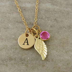 May include: A gold-toned necklace featuring a delicate chain with three charms: a circular disc engraved with the letter A, a detailed wing charm, and a small round pink gemstone set in a gold-toned bezel. The charms hang together from the chain.