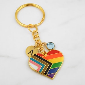 May include: Gold keyring with a rainbow heart charm, a small blue gemstone, and a round charm with the letter "A" engraved on it.